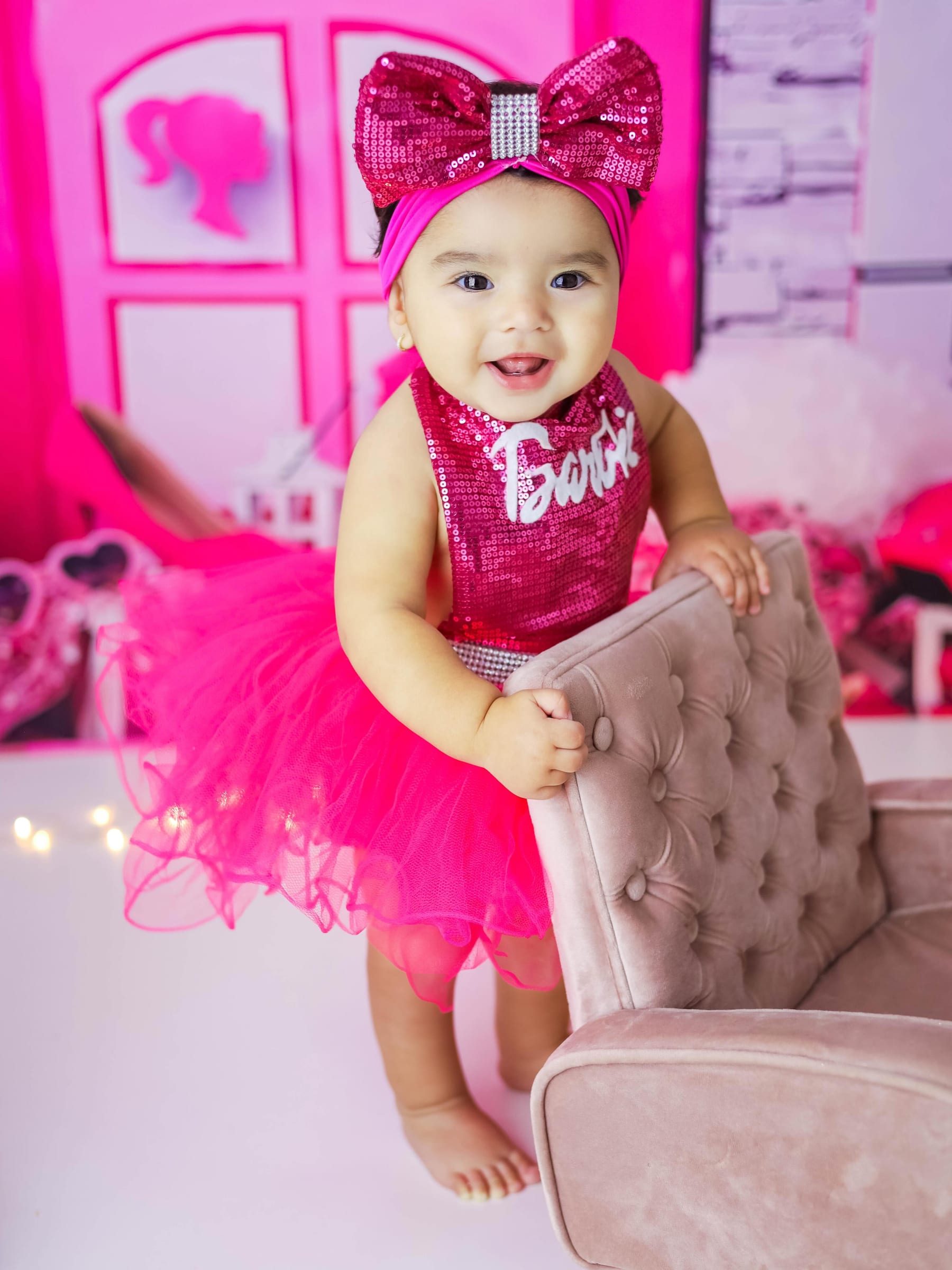 Bebê de vestido rosa sorrindo ao lado de uma poltrona