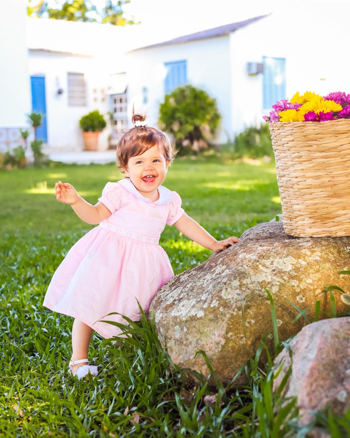 Menina de vestido rosa sorrindo em um jardim, ao lado de uma cesta de flores