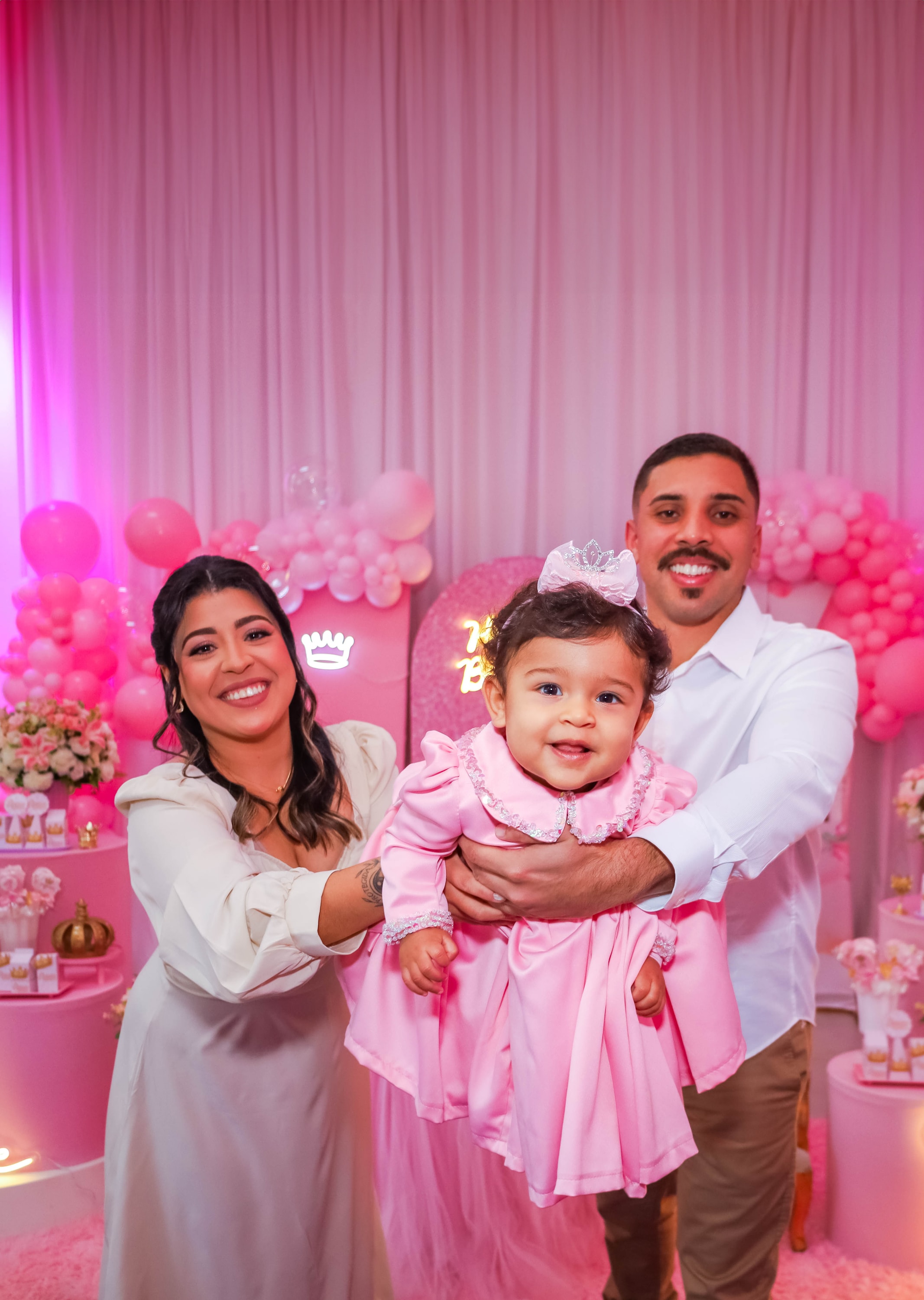 Pais sorridentes segurando a filha, vestida de rosa e coroa, durante festa de aniversário com decoração de balões