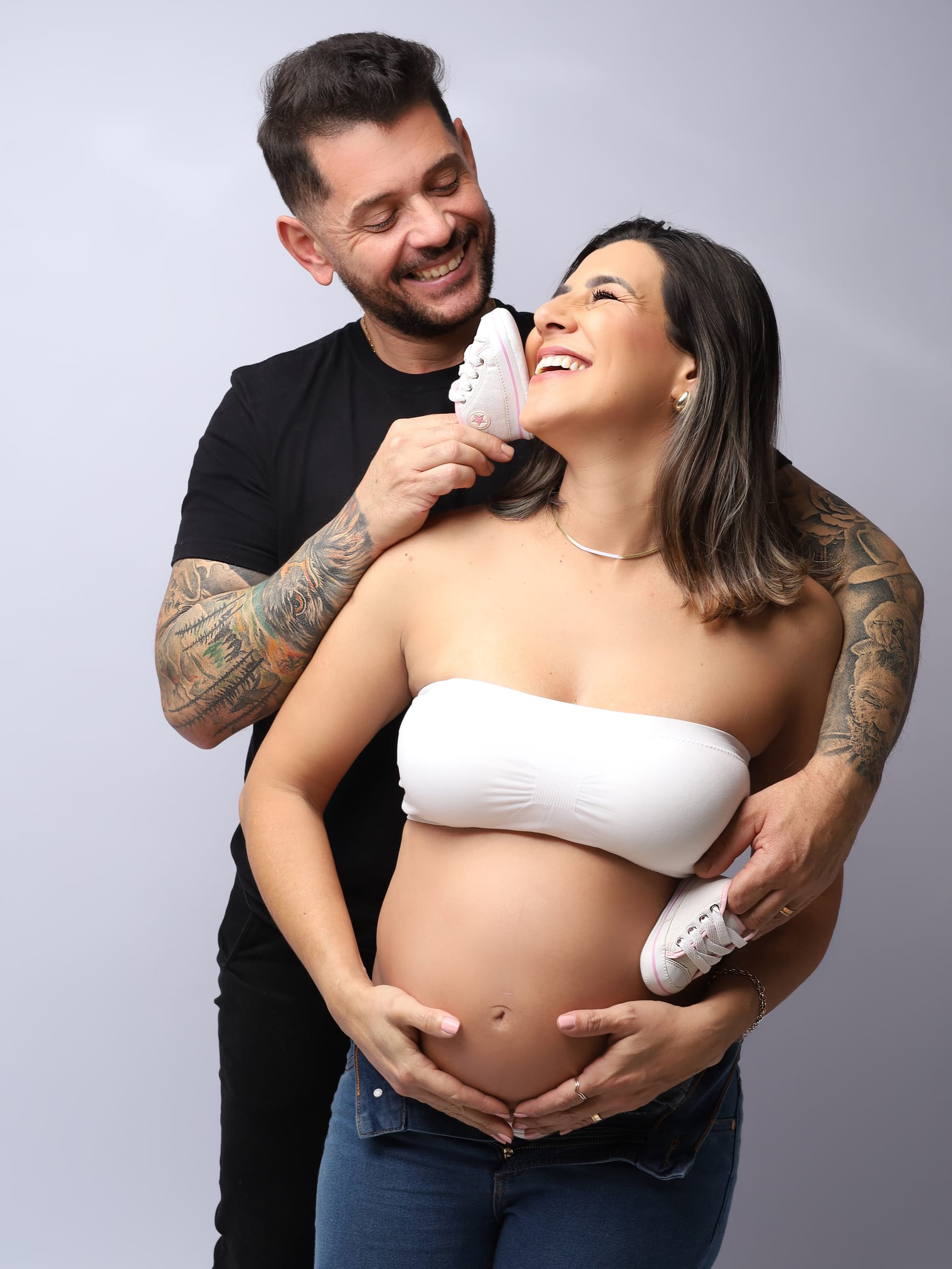 Casal sorrindo durante ensaio de gestante, abraçado e segurando sapatinhos de bebê