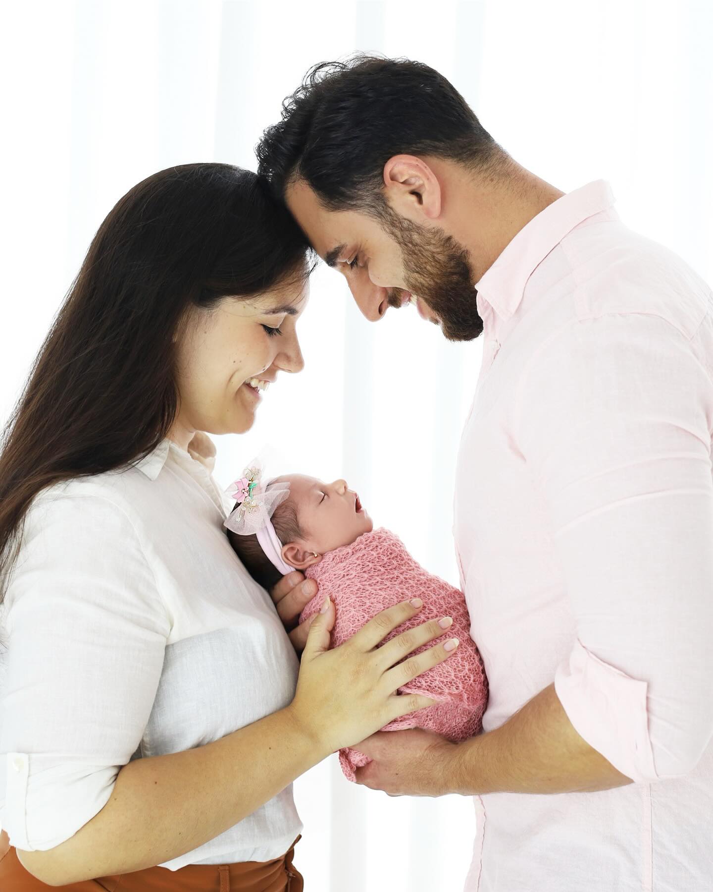 Pais com as testas coladas, sorrindo, segurando a filha recém-nascida envolvida em manta rosa